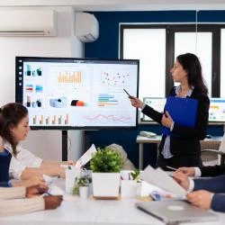 moça com terno preto fazendo apresentação com tela com planilhas, em um sala de reunião com várias pessoas